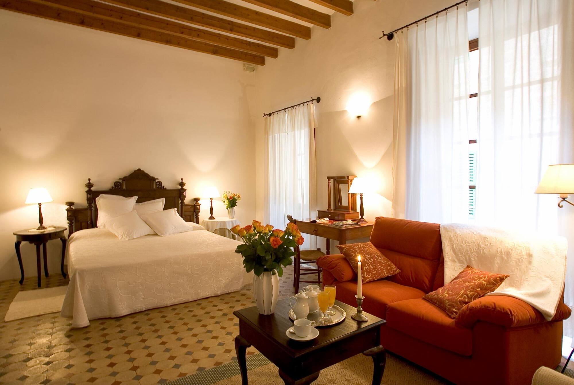 A suite with a white bed and a terracotta sofa under a beamed ceiling, on a patterned tile floor.
