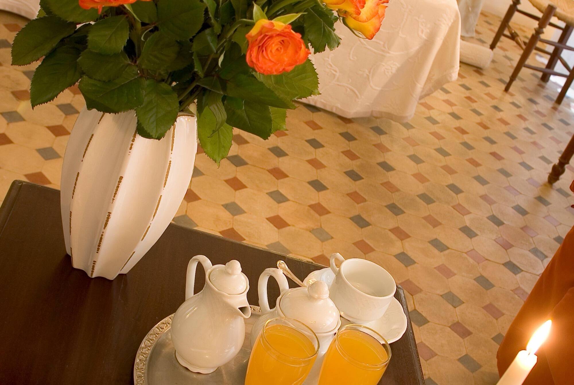 A breakfast tray of orange juice and a coffee service beside a vase of orange roses on a tiled floor.